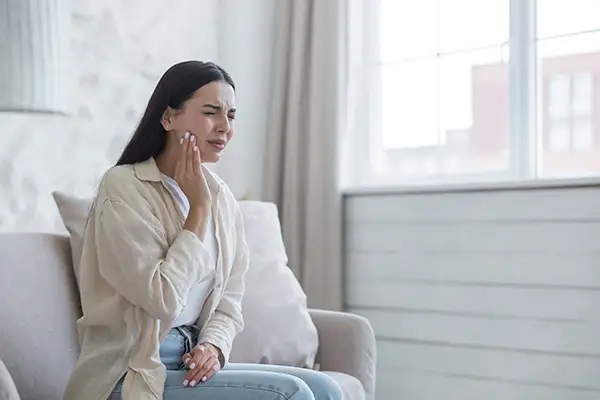 Young woman sitting on a couch, holding her cheek and wincing due to severe toothache, requiring emergency dental care at  Cascade Dental in Medford, OR
